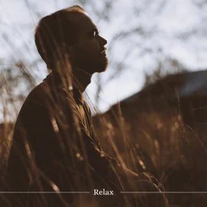 A side profile of a person sitting in a natural environment, bathed in warm, soft light. The image captures a serene moment, surrounded by tall grass and blurred natural scenery in the background.