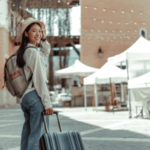 A smiling girl walking outdoors with a travel bag and a suitcase, ready to embark on a journey, with a scenic or urban backdrop